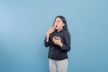 Mavi bir duvarın önünde duran bir kadın telefonuna bakıyor. Telefonundaki bir şey onu şaşırttı ya da şok etti.