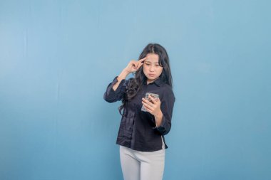 Bir kadın mavi bir duvarın önünde duruyor, elindeki tablete bakıyor. Derin düşüncelere dalmış, muhtemelen bir karar ya da sorun düşünüyor.