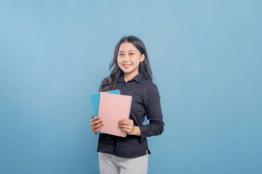 Bir kadın bir yığın kitap tutuyor ve gülümsüyor. O, siyah bir gömlek ve beyaz pantolon giyiyor. Güven ve pozitiflik kavramı