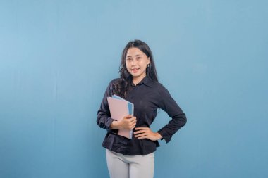 Bir kadın bir yığın kitap tutuyor ve gülümsüyor. O, siyah bir gömlek ve beyaz pantolon giyiyor. Güven ve pozitiflik kavramı