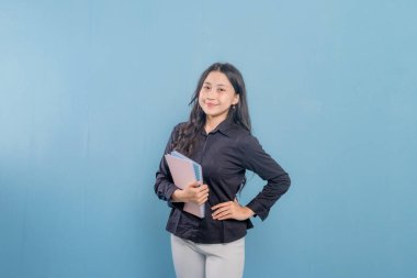 Bir kadın bir yığın kitap tutuyor ve gülümsüyor. O, siyah bir gömlek ve beyaz pantolon giyiyor. Güven ve pozitiflik kavramı