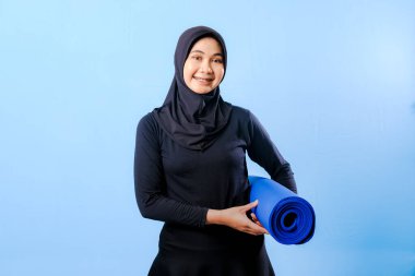 Siyah tesettürlü bir kadın mavi yoga minderi tutuyor. O gülümsüyor. Paspas sarılmış.