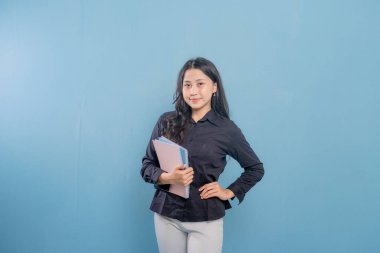 Bir kadın bir yığın kitap tutuyor ve gülümsüyor. O, siyah bir gömlek ve beyaz pantolon giyiyor. Güven ve pozitiflik kavramı