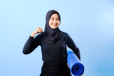 Siyah tesettürlü bir kadın mavi yoga minderi tutuyor. Gülümsüyor ve mutlu.