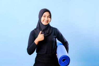 Siyah tesettürlü bir kadın mavi yoga minderi tutuyor. Başparmağıyla işaret veriyor.