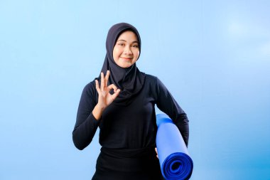 Siyah tesettürlü bir kadın mavi yoga minderi tutuyor. Başparmağıyla işaret veriyor.