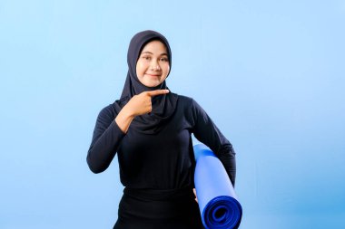 Siyah tesettürlü bir kadın bir şeyi işaret ediyor. Elinde mavi bir yoga minderi tutuyor. Paspas katlanır ve yere konur.
