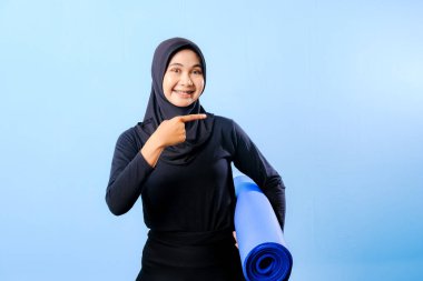 Siyah tesettürlü bir kadın bir şeyi işaret ediyor. Elinde mavi bir yoga minderi tutuyor. Paspas katlanır ve yere konur.