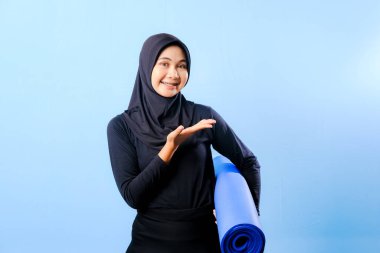 Siyah tesettürlü bir kadın bir şeyi işaret ediyor. Elinde mavi bir yoga minderi tutuyor. Paspas katlanır ve yere konur.