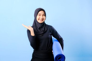 Siyah tesettürlü bir kadın bir şeyi işaret ediyor. Elinde mavi bir yoga minderi tutuyor. Paspas katlanır ve yere konur.