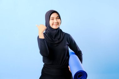 Siyah tesettürlü bir kadın bir şeyi işaret ediyor. Elinde mavi bir yoga minderi tutuyor. Paspas katlanır ve yere konur.