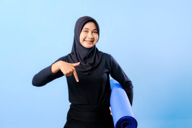 Siyah tesettürlü bir kadın bir şeyi işaret ediyor. Elinde mavi bir yoga minderi tutuyor. Paspas katlanır ve yere konur.