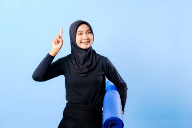 Siyah tesettürlü bir kadın bir şeyi işaret ediyor. Elinde mavi bir yoga minderi tutuyor. Paspas katlanır ve yere konur.