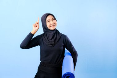 Siyah tesettürlü bir kadın bir şeyi işaret ediyor. Elinde mavi bir yoga minderi tutuyor. Paspas katlanır ve yere konur.