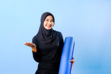 Siyah tesettürlü bir kadın bir şeyi işaret ediyor. Elinde mavi bir yoga minderi tutuyor. Paspas katlanır ve yere konur.