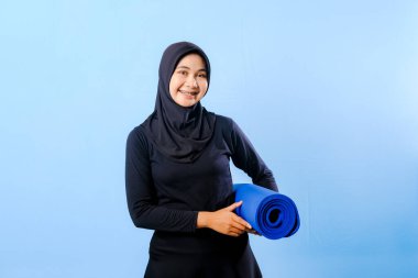 Siyah tesettürlü bir kadın mavi yoga minderi tutuyor. O gülümsüyor. Paspas sarılmış.