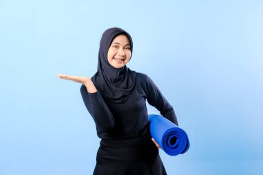 Siyah tesettürlü bir kadın bir şeyi işaret ediyor. Elinde mavi bir yoga minderi tutuyor. Paspas katlanır ve yere konur.