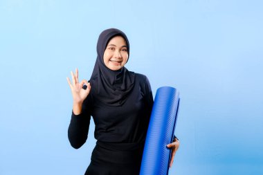 Siyah tesettürlü bir kadın mavi yoga minderi tutuyor. Başparmağıyla işaret veriyor.