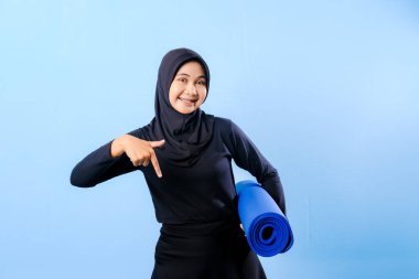 Siyah tesettürlü bir kadın bir şeyi işaret ediyor. Elinde mavi bir yoga minderi tutuyor. Paspas katlanır ve yere konur.