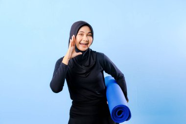 Siyah başörtüsü ve mavi pantolon giyen bir kadın eğiliyor. Bağırarak ya da bir şey anons ederek bir jest yapıyor. Elinde mavi bir yoga minderi var.