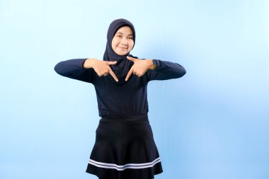 Siyah etek ve siyah başörtüsü takan bir kadın. Bir şeyi işaret ediyor. Gülümsüyor.