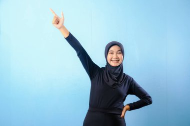 Siyah etek ve siyah başörtüsü takan bir kadın. Bir şeyi işaret ediyor. Gülümsüyor.