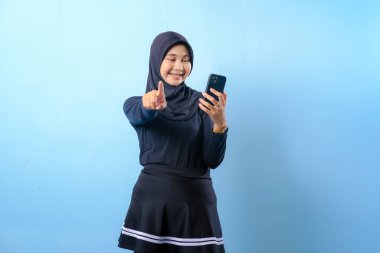 Siyah tesettürlü bir kadın cep telefonu tutuyor ve kamerayı işaret ediyor. Gülümsüyor ve mutlu.