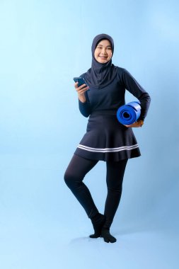 Siyah etekli ve siyah pantolonlu bir kadın mavi yoga minderi tutuyor. Cep telefonuna bakıyor.