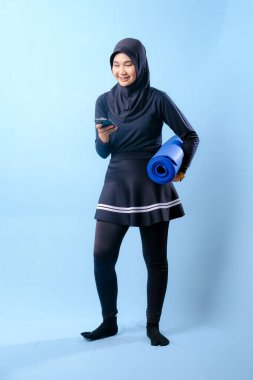 Siyah etekli ve siyah pantolonlu bir kadın mavi yoga minderi tutuyor. Cep telefonuna bakıyor.