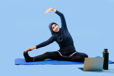 Bir kadın laptopu ve su şişesi olan bir minderde yoga yapıyor. Gülümsüyor ve antrenmanından zevk alıyor.