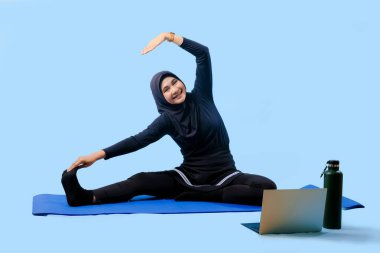 Bir kadın laptopu ve su şişesi olan bir minderde yoga yapıyor. Gülümsüyor ve antrenmanından zevk alıyor.