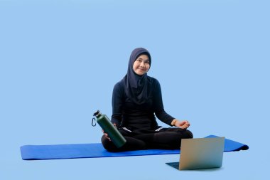 Bir kadın yoga minderinde dizüstü bilgisayarı ve su şişesiyle oturuyor. Gülümsüyor ve eğleniyor.