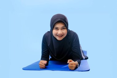 Siyah tesettürlü bir kadın mavi bir paspasın üzerinde yatıyor. Gülümsüyor ve eğleniyor.