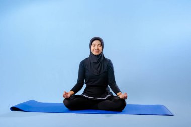 Siyah tesettürlü bir kadın mavi bir paspasın üzerinde oturuyor. Gülümsüyor ve rahatlamış durumda.