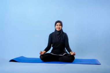 Siyah tesettürlü bir kadın mavi bir paspasın üzerinde oturuyor. Gülümsüyor ve rahatlamış durumda.