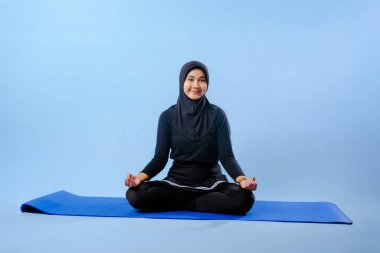 Siyah tesettürlü bir kadın mavi bir paspasın üzerinde oturuyor. Gülümsüyor ve rahatlamış durumda.