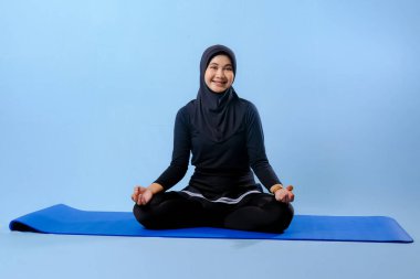 Siyah tesettürlü bir kadın mavi bir paspasın üzerinde oturuyor. Gülümsüyor ve rahatlamış durumda.