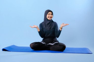 Siyah tesettürlü bir kadın mavi bir paspasın üzerinde oturuyor. Gülümsüyor ve rahatlamış durumda.