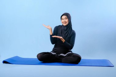 Siyah tesettürlü bir kadın mavi bir paspasın üzerinde oturuyor. Gülümsüyor ve rahatlamış durumda.