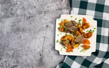 Kare tabakta koyu gri bir zeminde fırında patates ve havuçla pişirilmiş ördek yahnisi. Üst manzara, düz uzanma