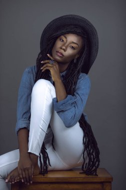 Stüdyoda gri arka planda, renkli mücevherler, yaz şapkası, kot pantolon ve kırmızı pantolon pantolon askıları takan, uzun saçlı ve güzel makyajlı genç bir kadının portresi..