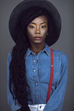 Uzun rastalı, güzel makyajlı, gri arka planda, yaz şapkası, kot gömlek ve kırmızı pantolon askısı takan genç bir siyahi kadının portresi..