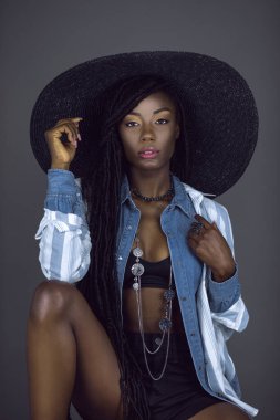 Uzun saçlı, uzun saçlı, güzel makyajlı, nemli dudakları olan, gri arka planda bir stüdyoda gri bir şapka ve takı takan genç ve seksi bir kadının portresi..