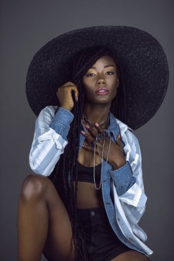 Şehvetli, genç, siyah bir kadının portresi. Uzun saçlı, güzel makyajlı, nemli dudakları var. Gri arka planda bir stüdyoda kendi kendine poz veriyor..