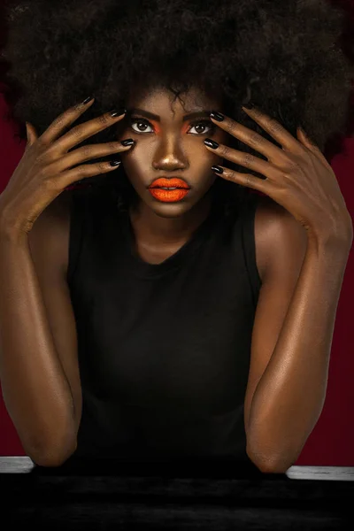 Portrait of a flirtatious young black female with long black hair, popping orange lip stick, perfectly manicured nails is wearing a big afro & leaning on a table in front of a dark red background.