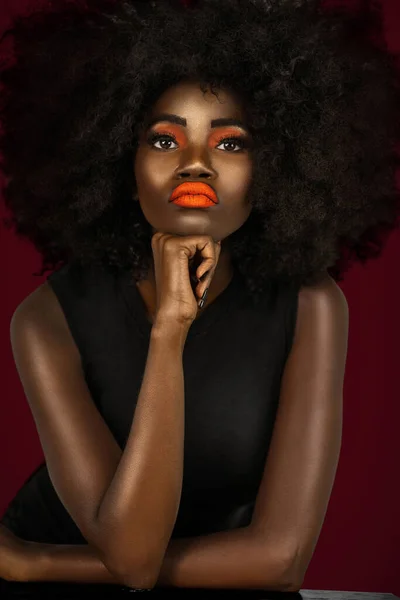 一个有着一头长长的黑头发 长着桔黄色唇膏 修剪得完美的指甲的宁静的年轻黑人女性的画像 穿着一件大大的非洲衣服 靠着桌子 面对着漆黑的红色背景 — 图库照片