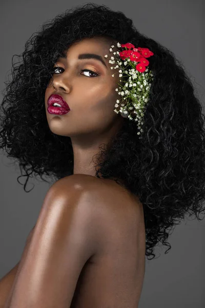 A portrait of a sexy young black female with curly long black hair, beautiful makeup and red lips posing by herself in a studio with grey background wearing flowers in her hair.