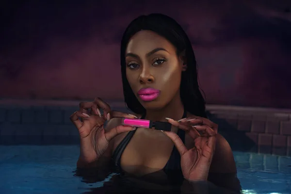 A portrait of a sexy young black female with long black hair, beautiful makeup, popping pink lip stick & perfectly manicured nails posing by herself inside a pool holding a pink lip stick.