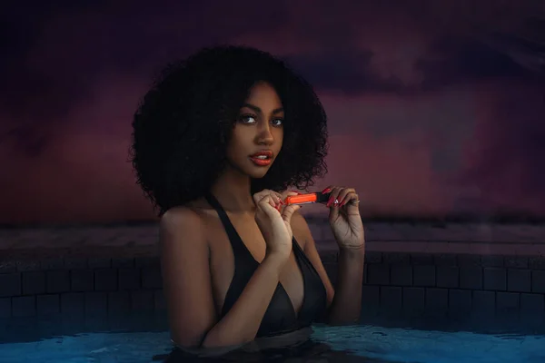 A gorgeous young black female with beautiful makeup & long black curly hair posing by herself inside a pool wearing a bikini top and holding an orange lipstick in her hands.