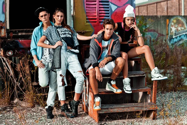 A portrait of an interracial group of 4 young adults sitting on wooden steps in front of an open graffiti sprayed container on a vacant lot during a pleasant summer day wearing cool urban outfits.